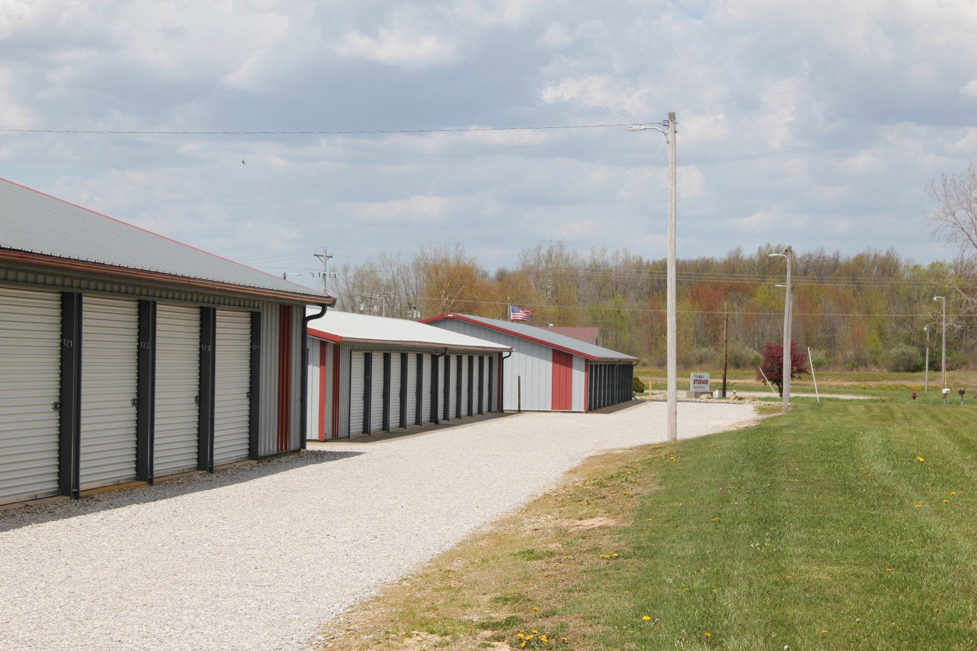 Storage units along a gravel road, red and gray buildings, green lawn, cloudy sky.