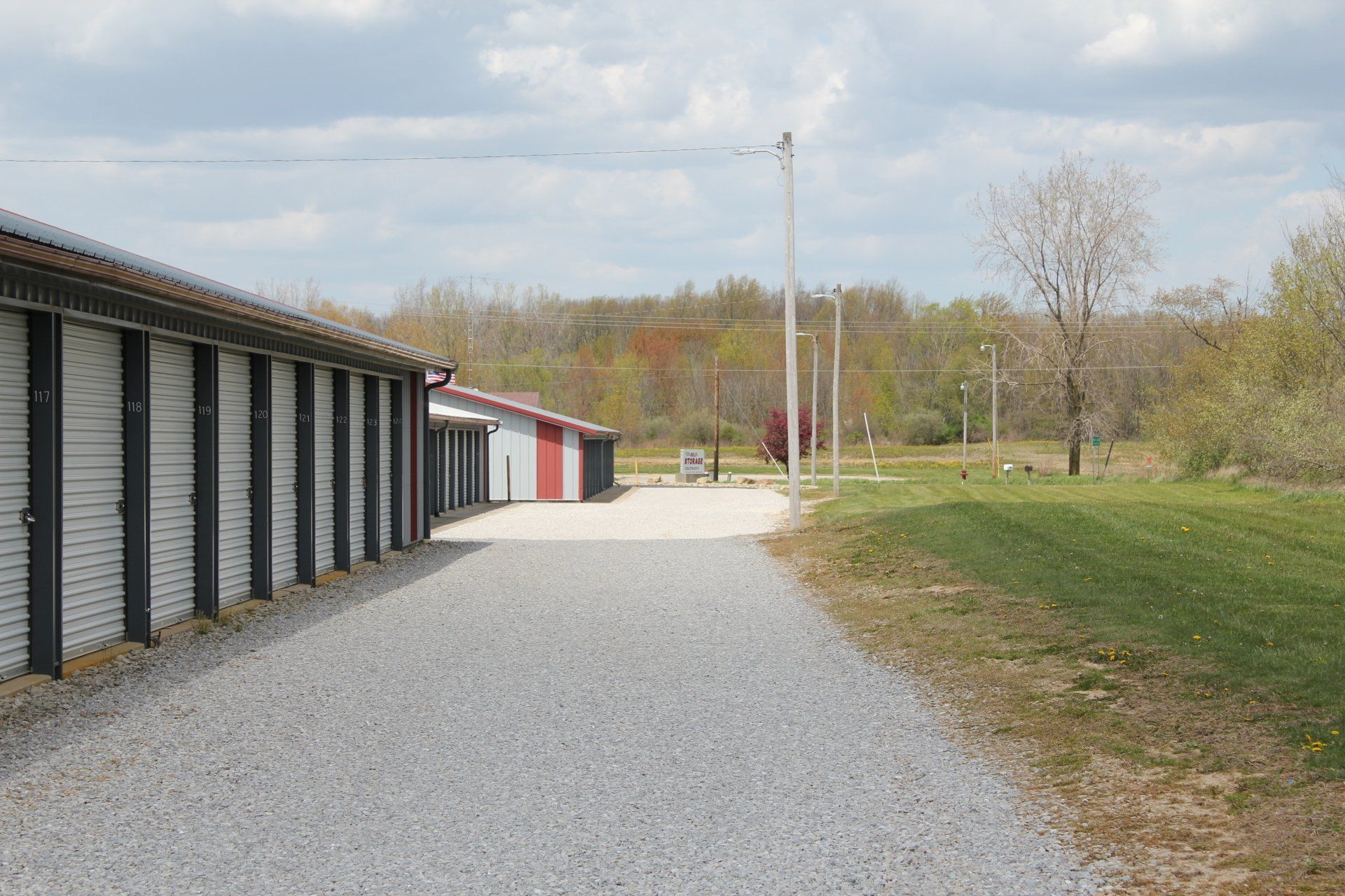 Exterior of storage units with gravel driveway and wooded area. Cloudy sky overhead.