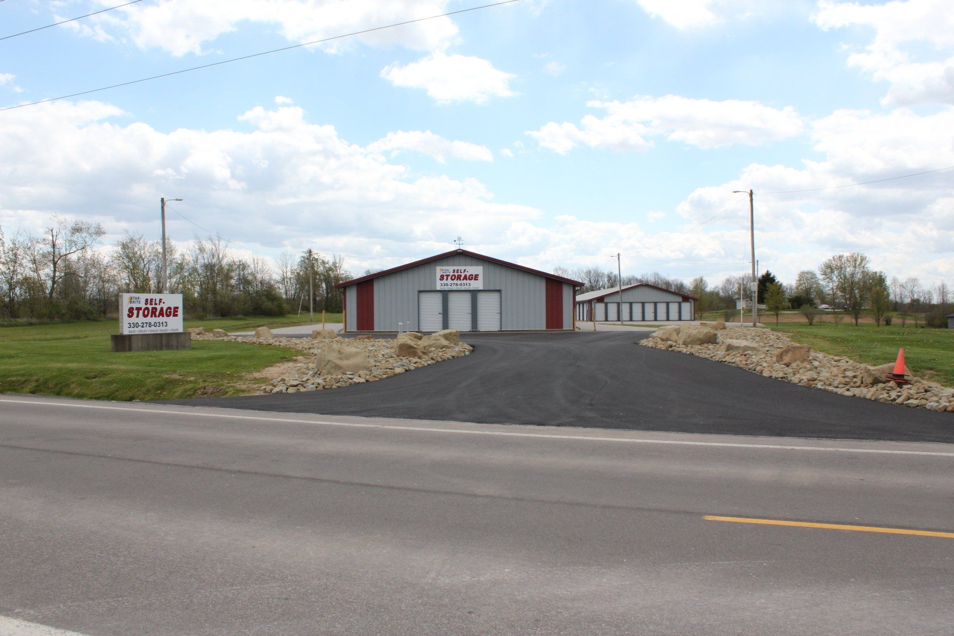 Storage facility with gray and red metal buildings, asphalt drive, and sign on a grassy lot.