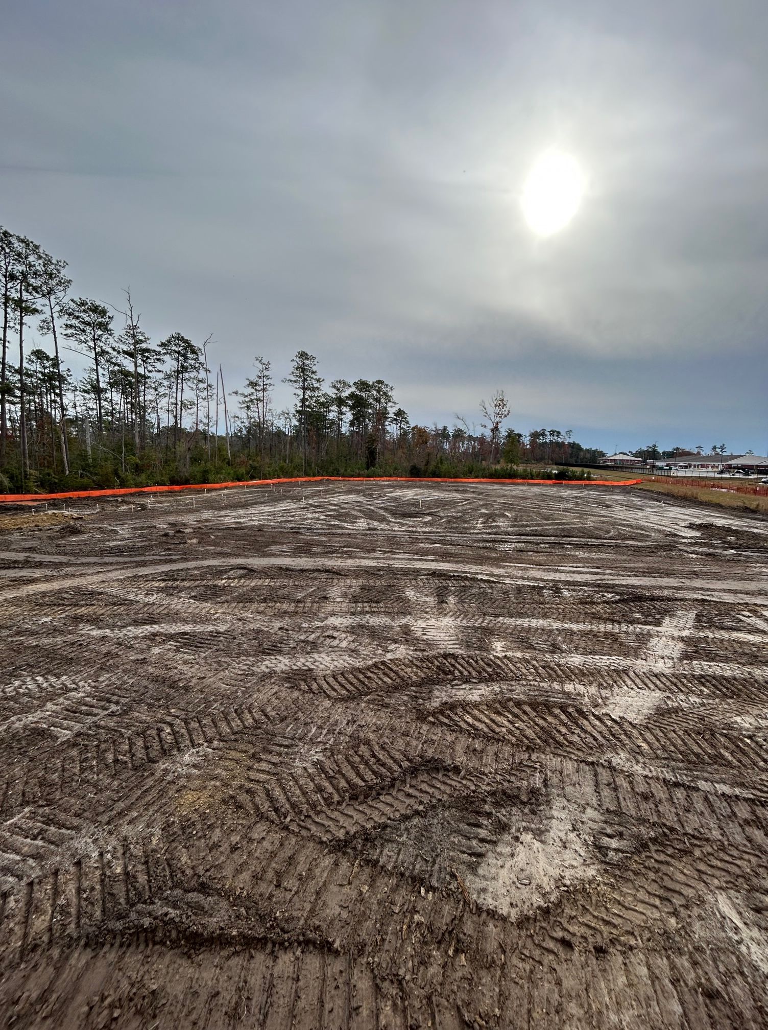 A large dirt field with trees in the background and the sun shining through the clouds.
