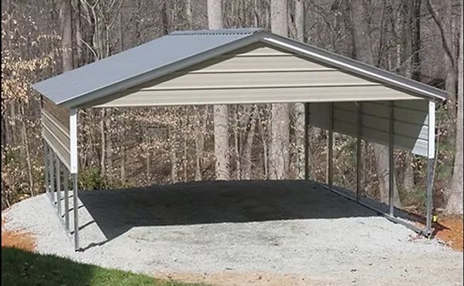 A metal carport with a tan siding and gray roof, gravel base, set against a backdrop of trees.