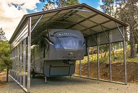 RV parked under a metal carport in a wooded setting.
