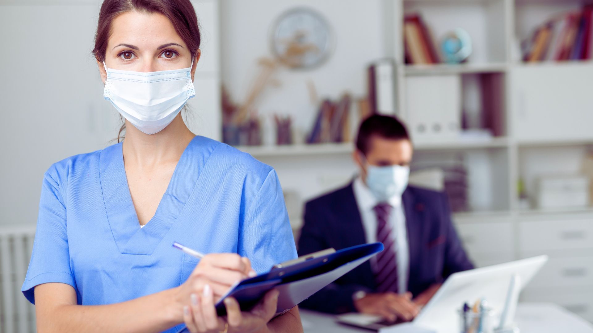 Un'infermiera che indossa una maschera tiene in mano un blocco appunti e ci scrive sopra.