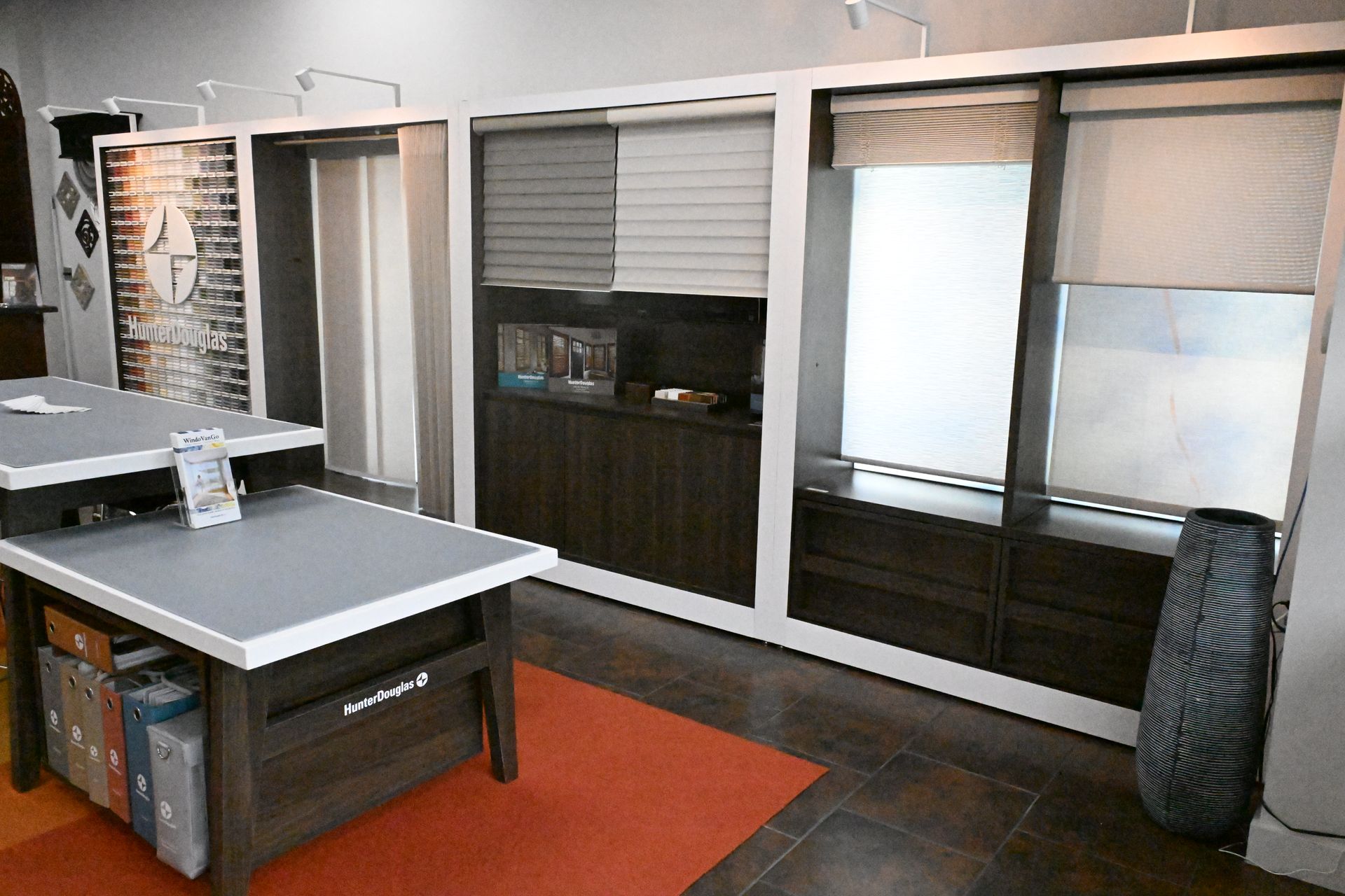 A showroom featuring window blinds displays and samples, with a large work table in the foreground on an orange rug.