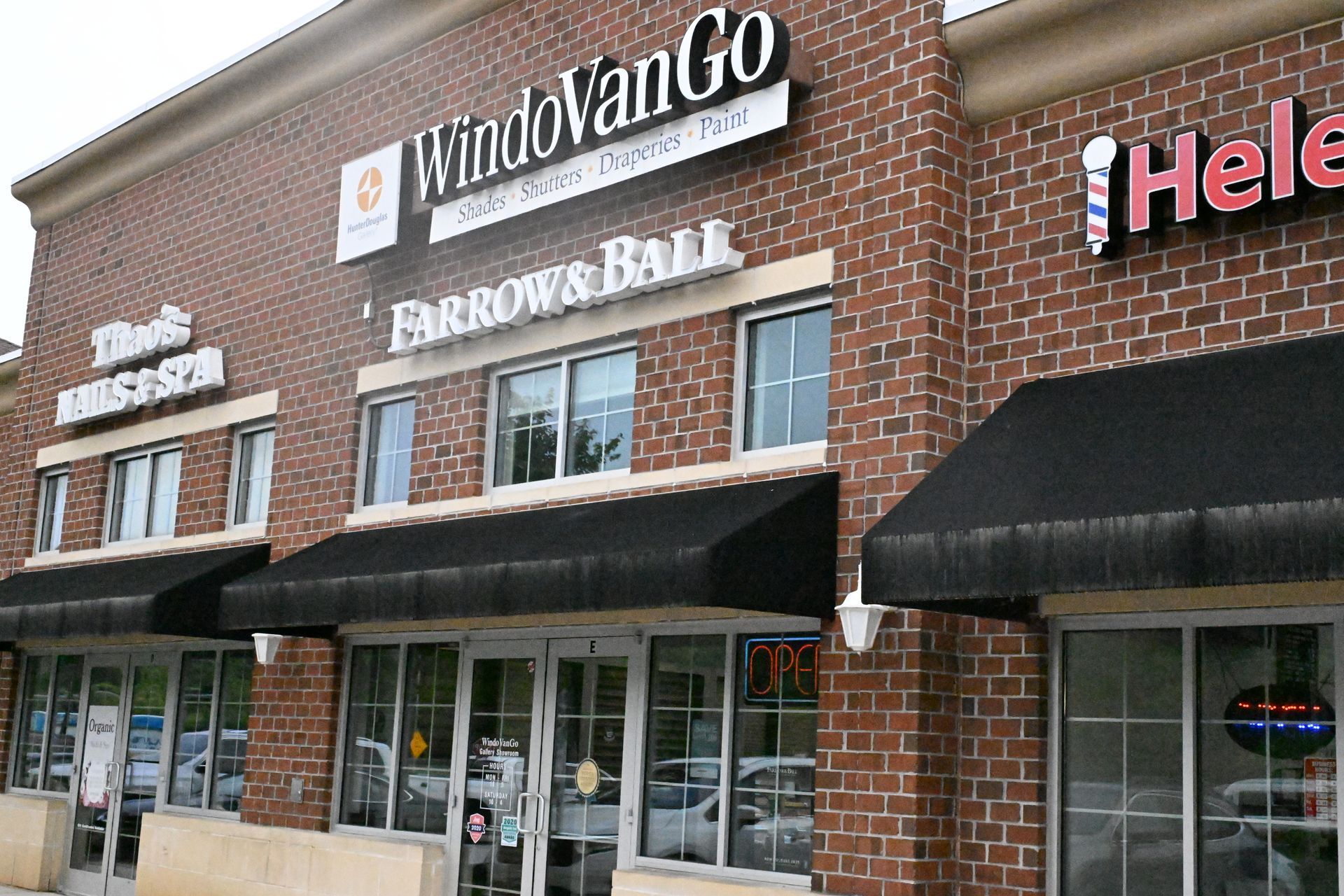 A brick retail strip center with signage for WindoVanGo, Farrow & Ball, and a barber shop under black fabric awnings.