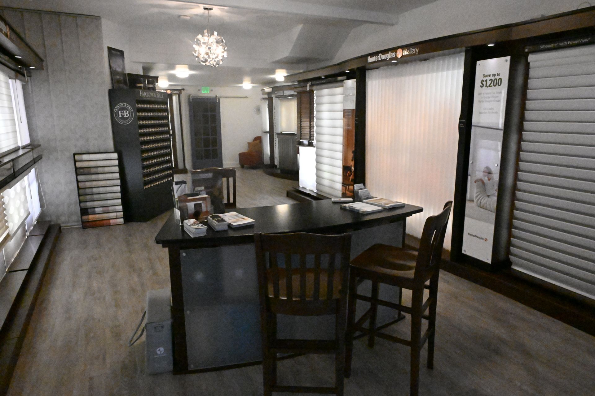 A showroom for window blinds and shades featuring displays, a dark desk with two chairs, and light wood-look flooring.