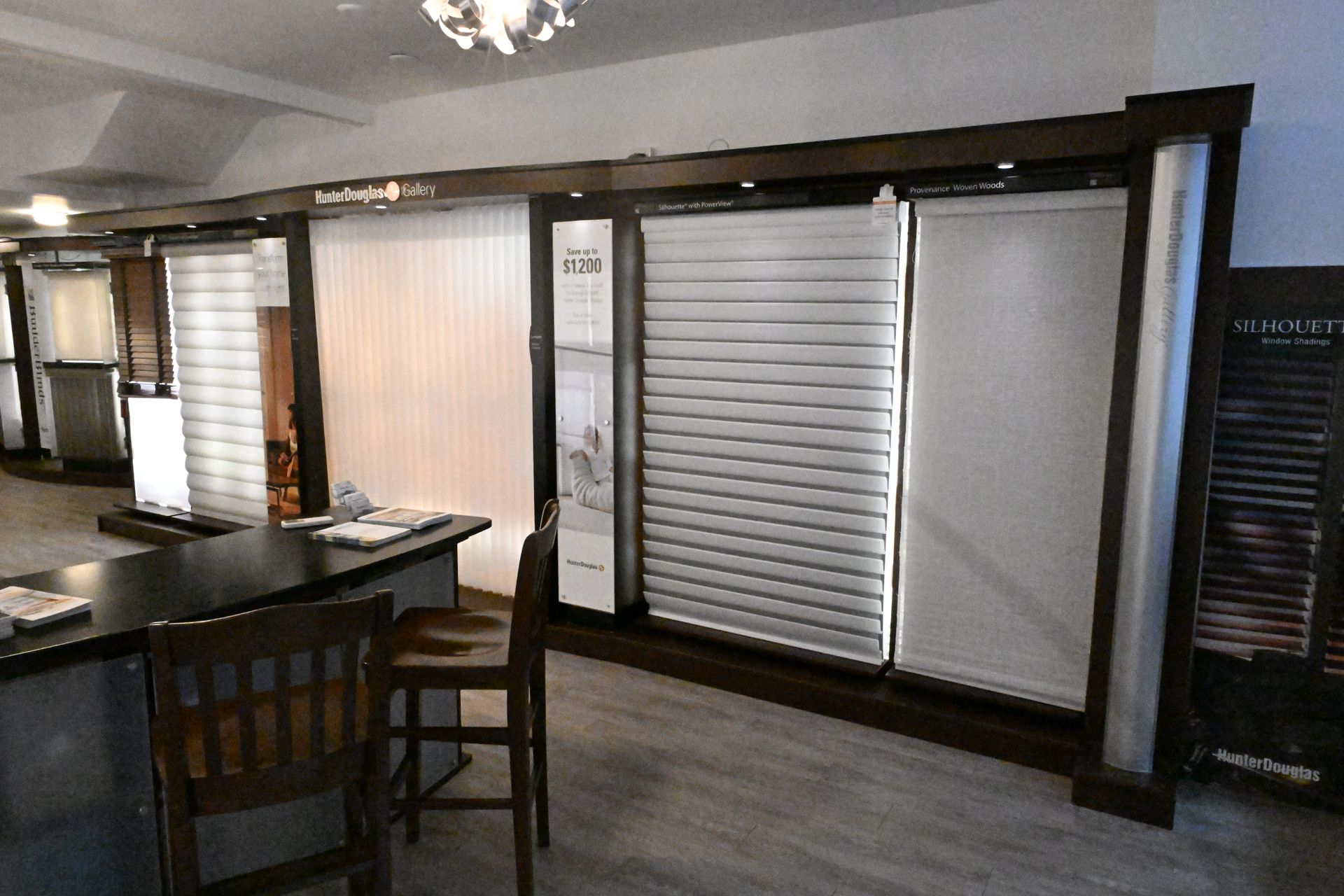 A showroom featuring various styles of window blinds and shades displayed in dark wooden frames, with chairs and a desk.