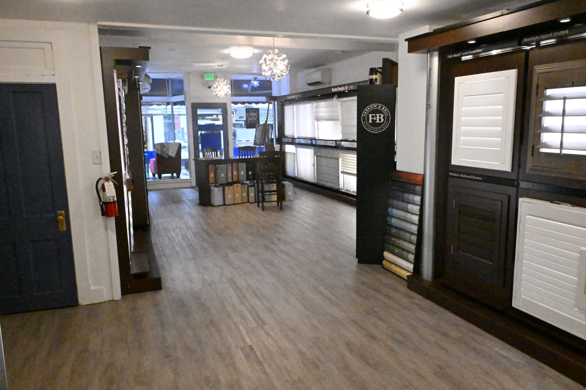 A showroom featuring various window shutter and blind displays, with a dark blue door on the left and wood-look flooring.