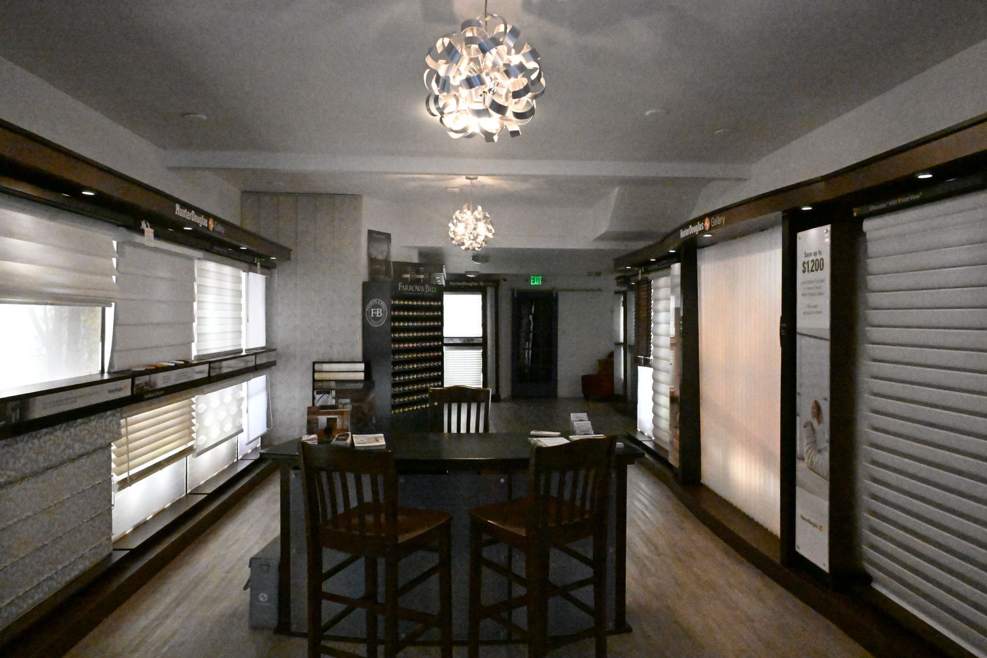 A window treatment showroom with displays of various blinds and shades, a central table, two chairs, and light fixtures.
