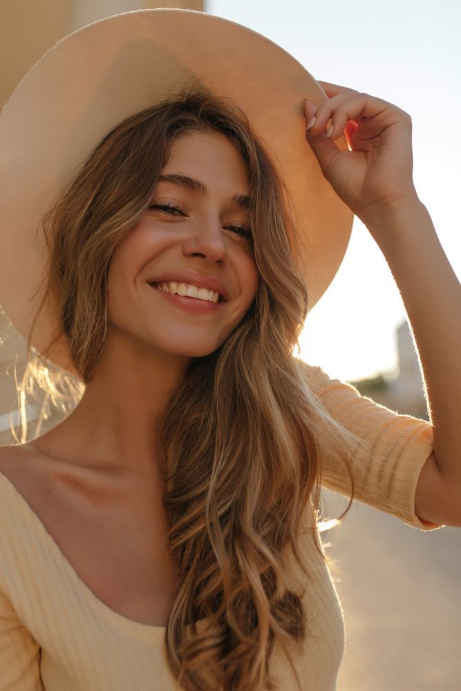Woman in a sun hat smiling, holding the brim with sunlight illuminating her face and hair.