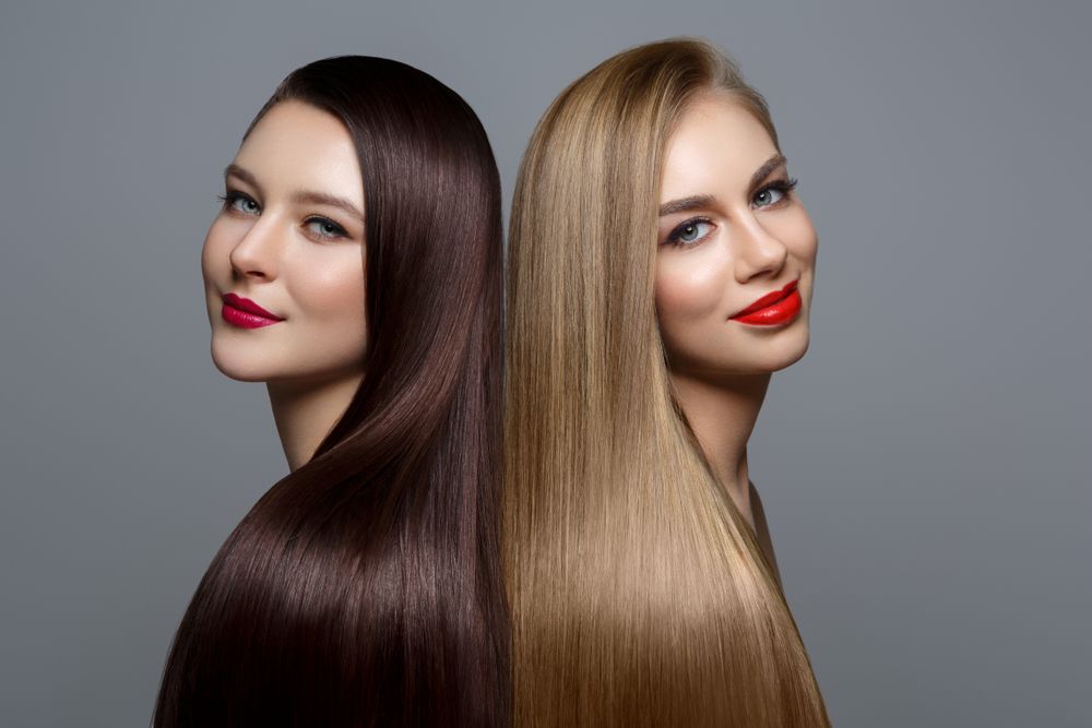 Two women with long, straight hair of contrasting dark brown and blonde colors, smiling, back-to-back.