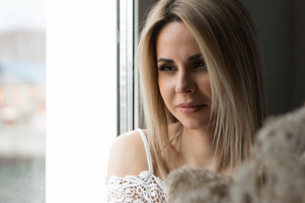 Blonde woman looking out a window; wearing white lace top, soft focus, natural light.