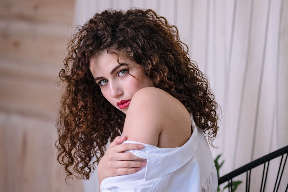 Woman with curly brown hair, wearing a white off-the-shoulder shirt, looking over her shoulder.
