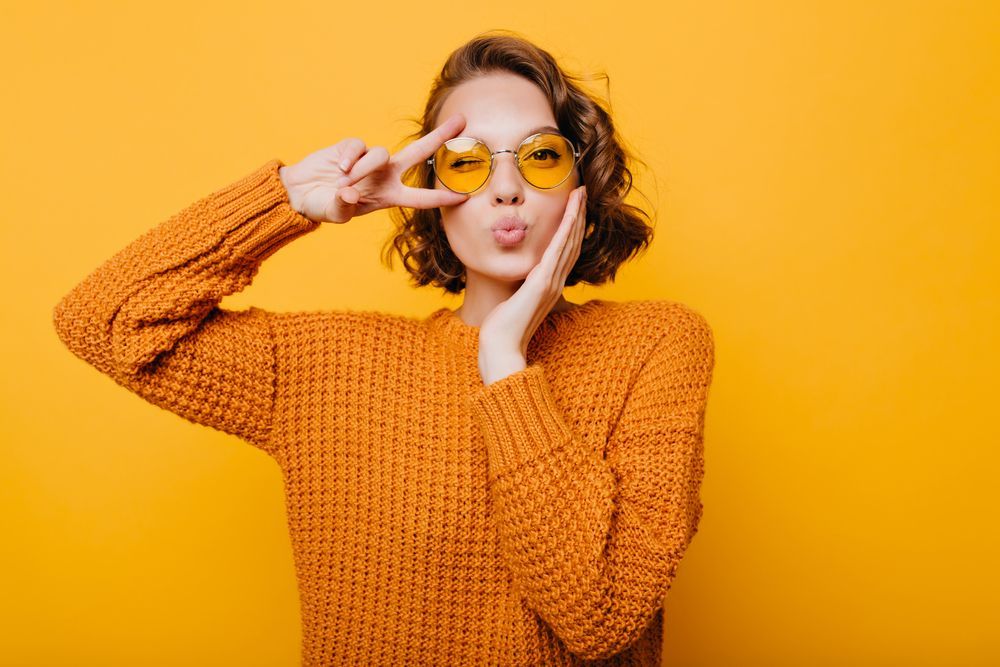 Woman in orange sweater and sunglasses making a peace sign with one hand, pouting, with yellow background.