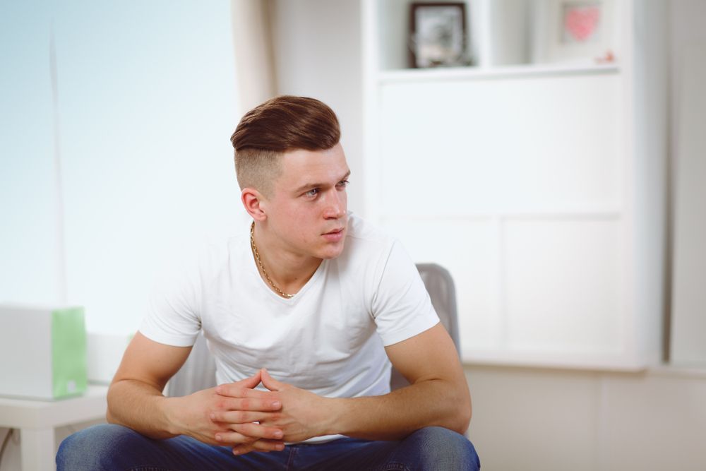 Man sitting, looking to the side, wearing a white t-shirt and jeans. Hands clasped, indoors with neutral background.