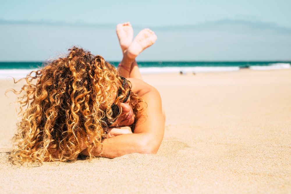 Woman with curly hair sunbathing on a sandy beach, feet up, ocean in background.