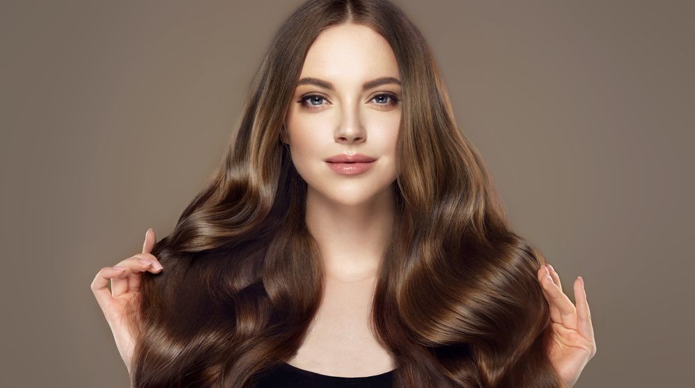 Woman with long, wavy, shiny brown hair, holding some strands, smiling, against a neutral background.