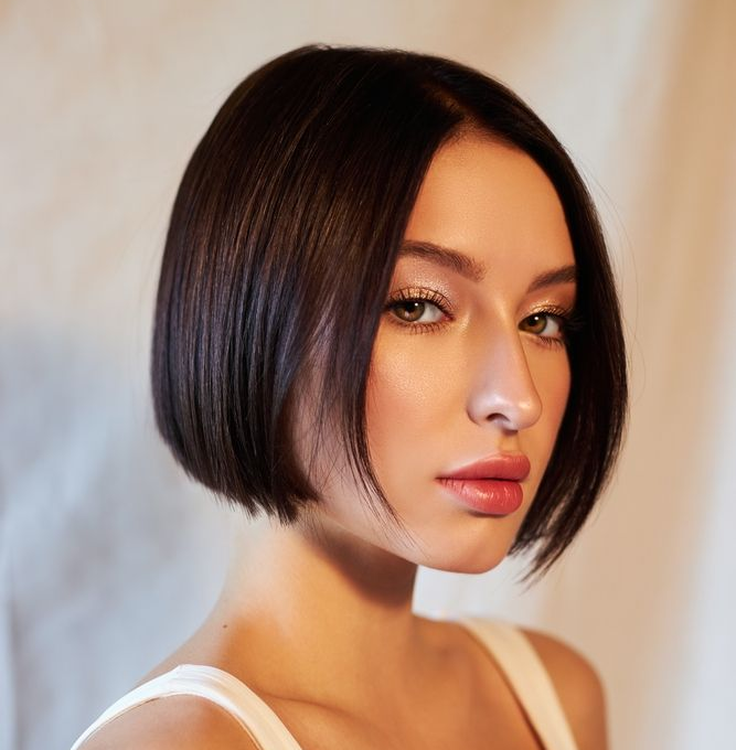 Woman with short dark hair, wearing a white tank top, looking towards the camera.