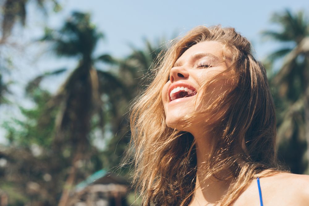 Woman laughing with eyes closed, backlit by sunlight, palm trees in background.