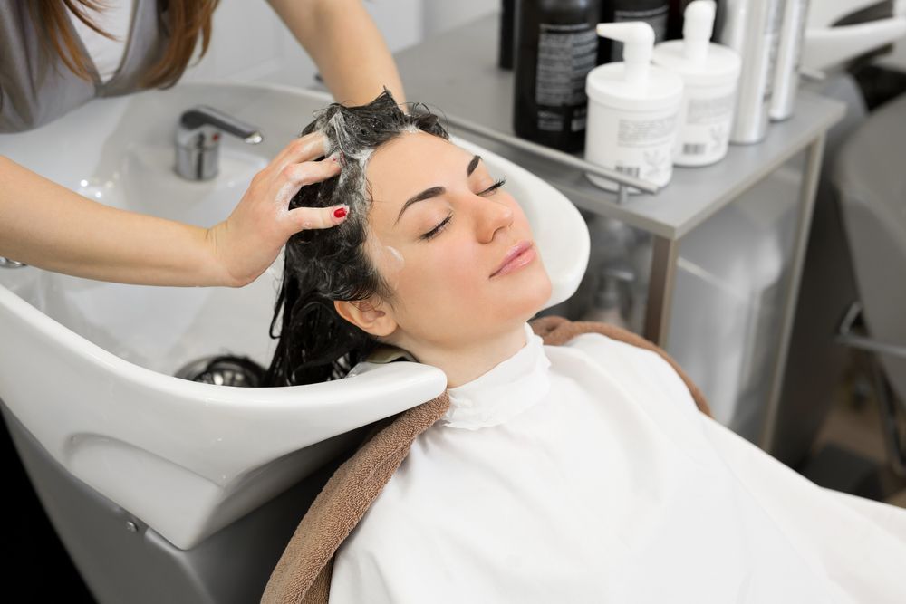 Woman having her hair washed at a salon, eyes closed, white cape, shampoo in hair.