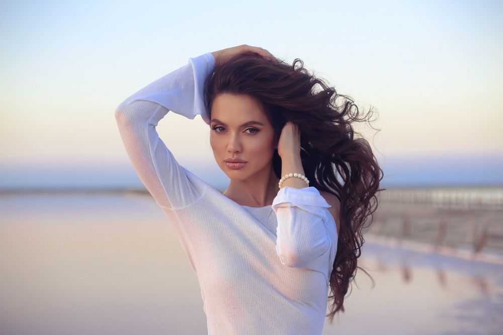 Woman with long, wavy brown hair poses outdoors, hands in hair, wearing white top; soft sunset in background.