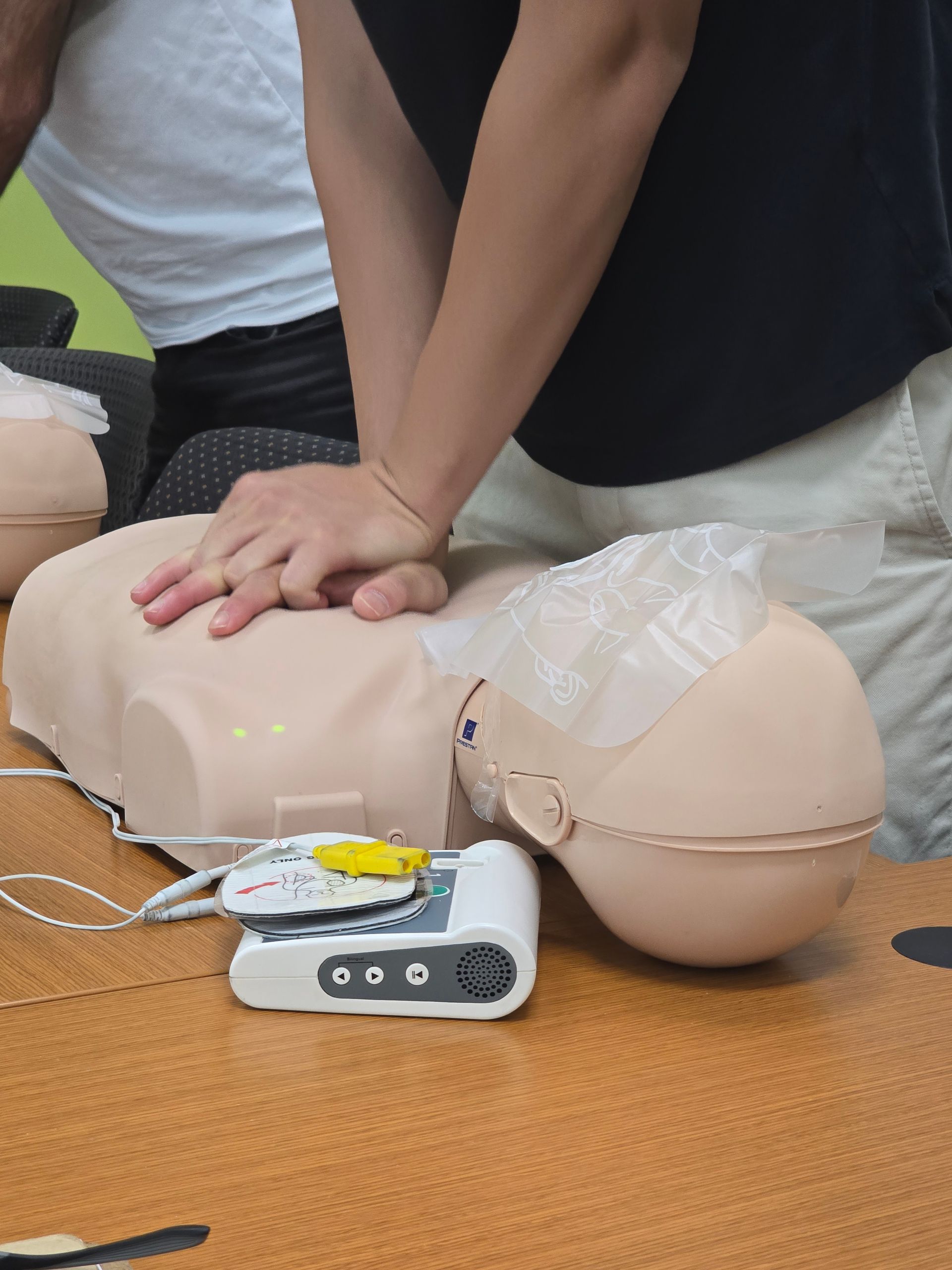 Hands performing CPR on a training mannequin, with an automated external defibrillator (AED) nearby.