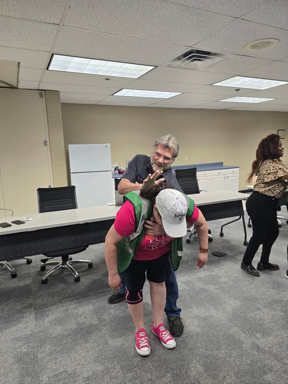 Man assisting person with a white hat, indoors. Person in a red shirt is being supported by the man. Others in the background.
