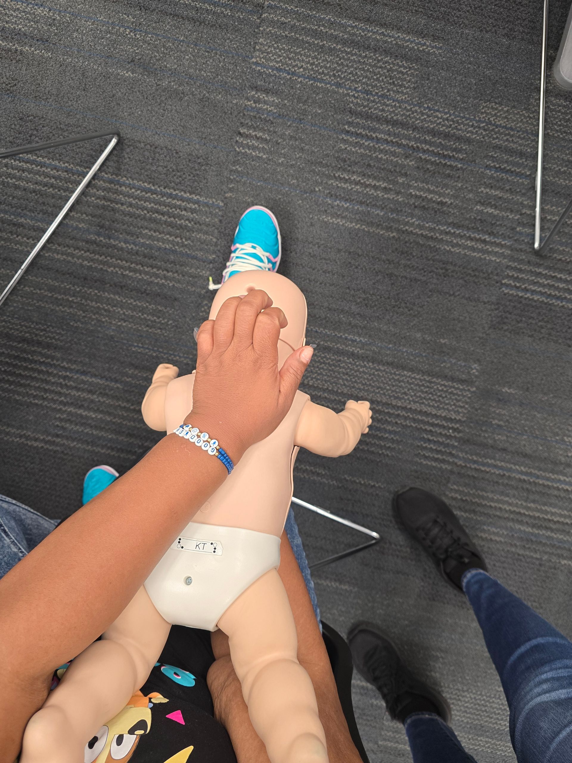 Person performing CPR on a baby mannequin, indoors. Blue shoes visible.