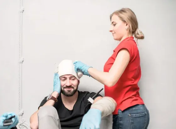 Woman in red shirt bandages man's head, both wearing gloves. Arm is bandaged; they are indoors.