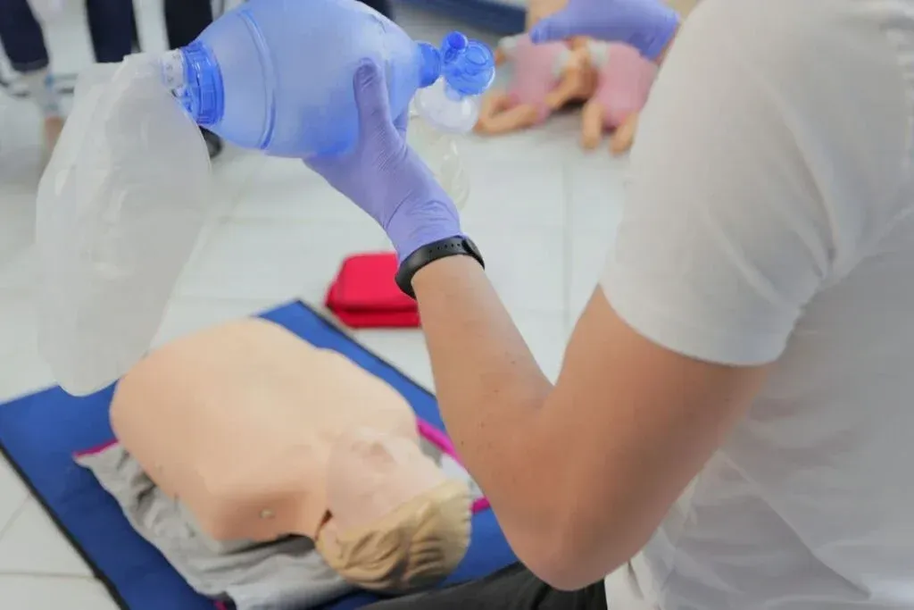 Person in gloves uses a bag valve mask on a CPR mannequin, likely in a training setting.