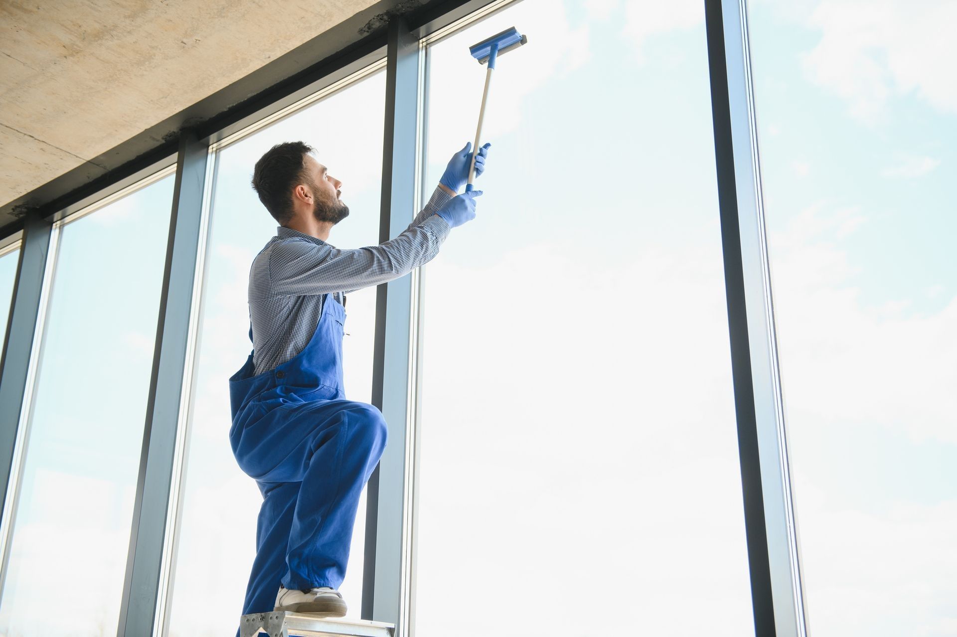 Man in blue overalls on a ladder, cleaning a large window with a squeegee. Indoor shot.