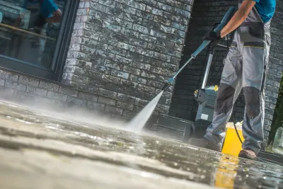 Person power washing a brick wall and paved surface, removing dirt with a high-pressure water stream.