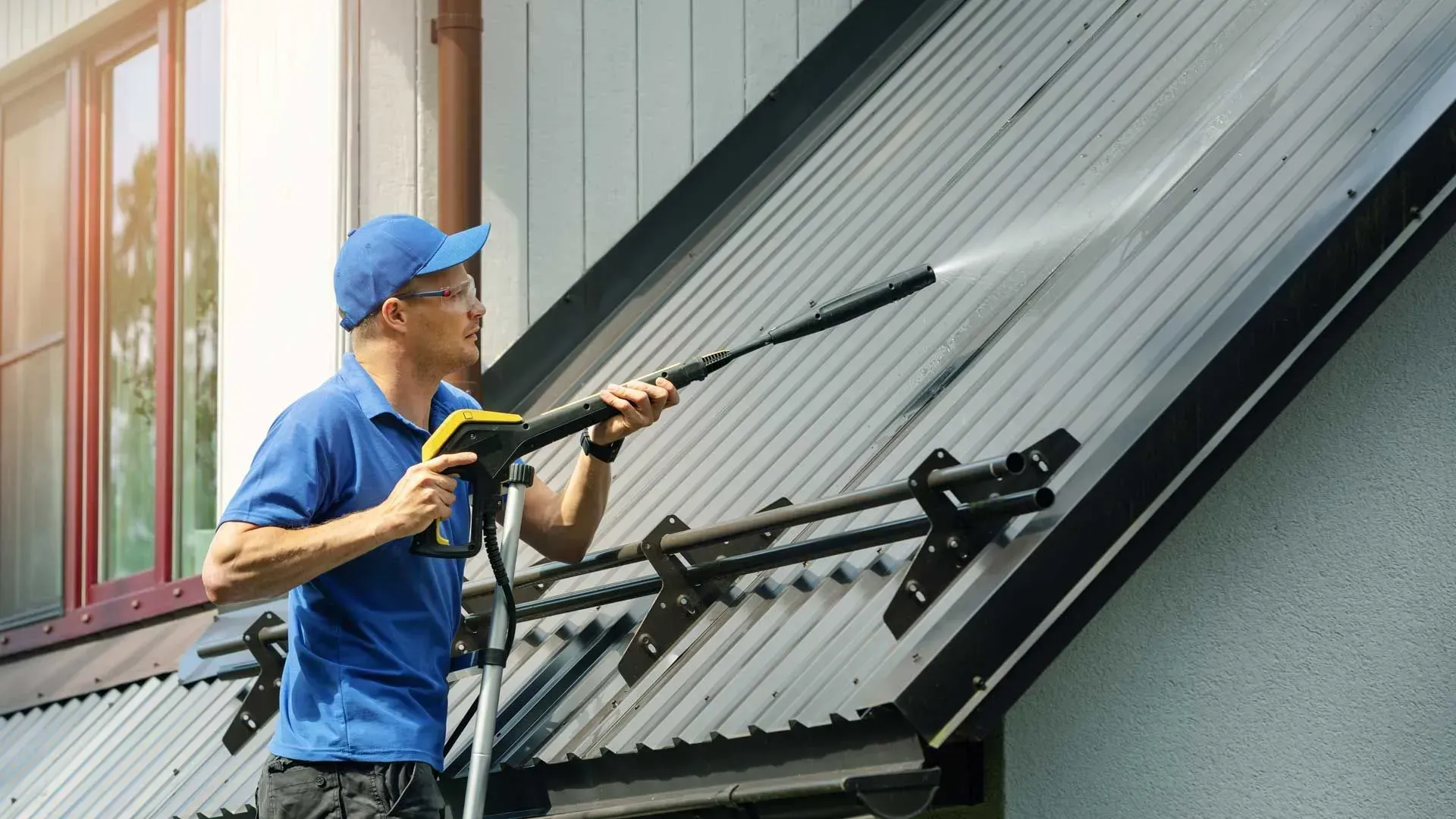 Man power washing a metal roof on a sunny day; he wears a blue cap and shirt.