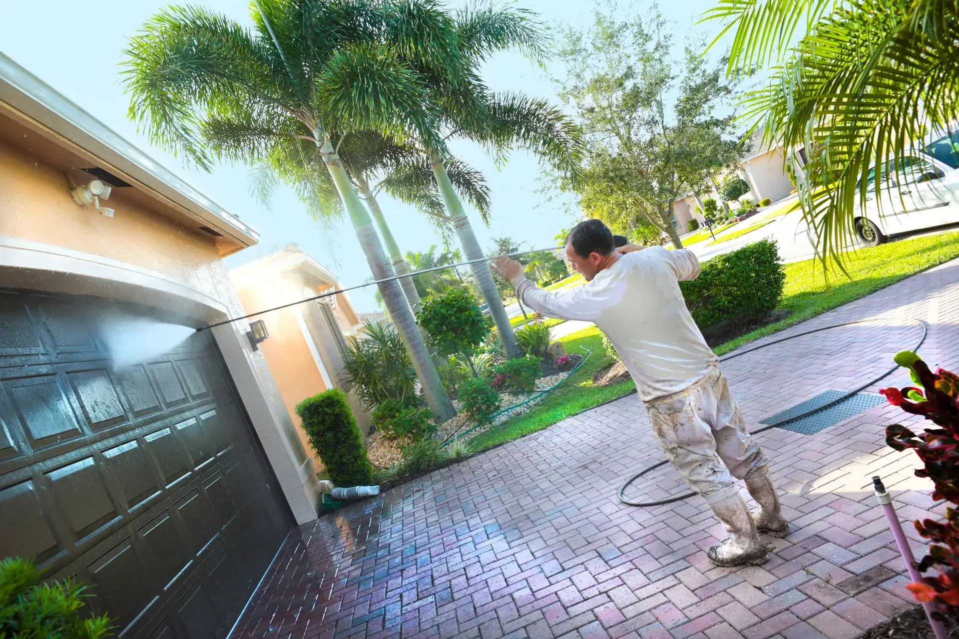 Man pressure washing a brick driveway in front of a garage. Bright sun and tropical landscaping in the background.