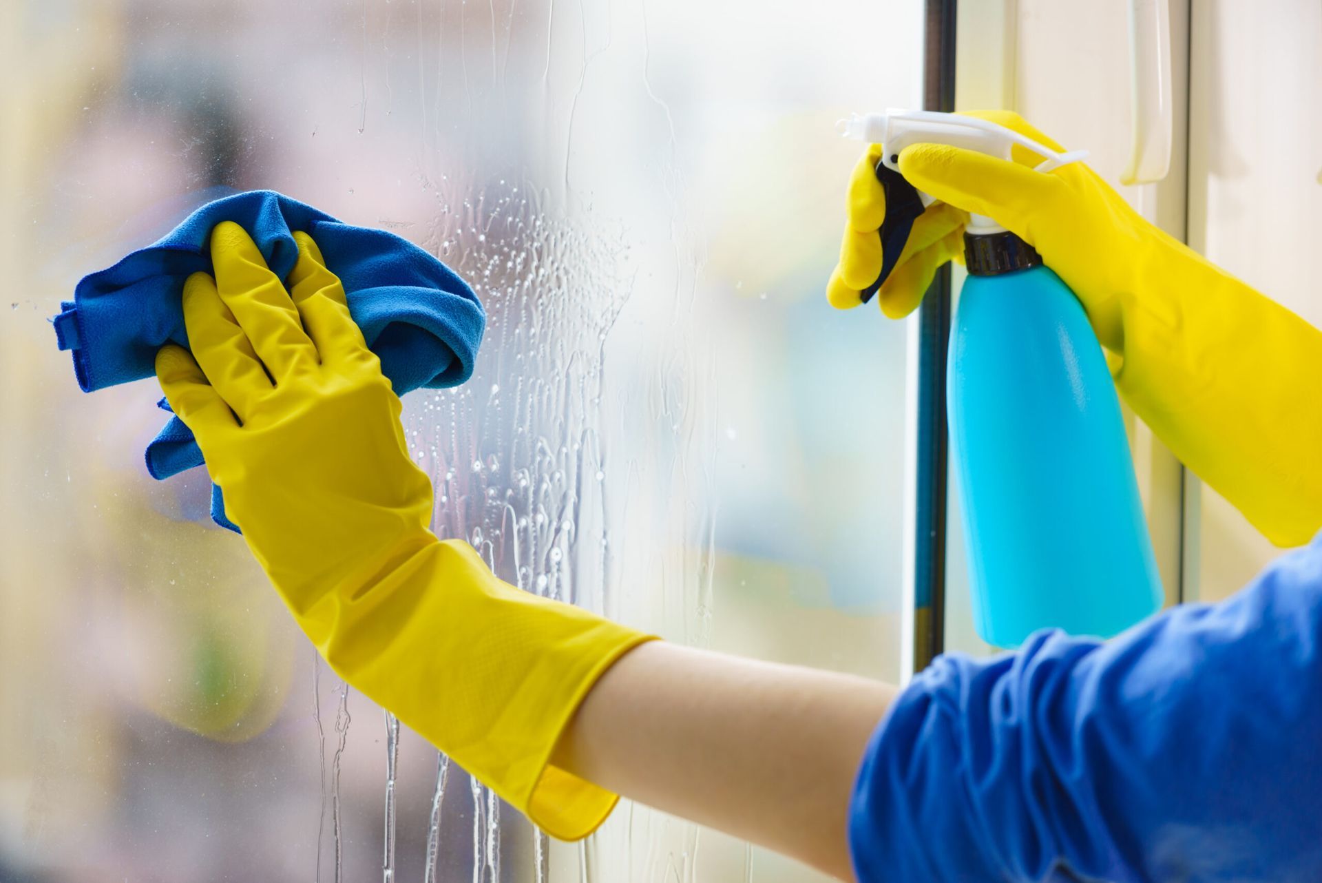 Person wearing yellow gloves cleaning a window with a blue cloth and spray bottle.