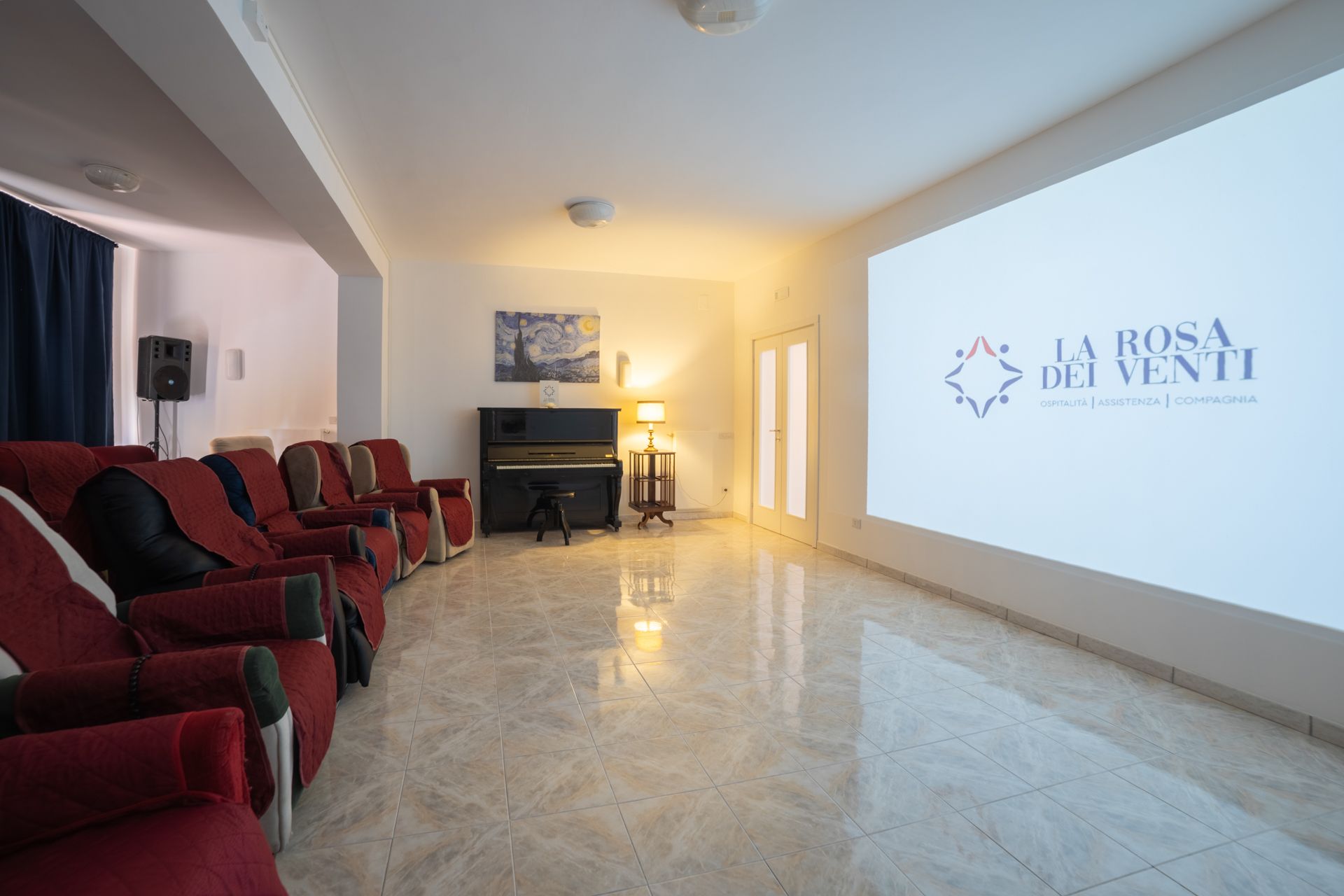 Una sala cinematografica accogliente con file di poltrone rosse, un pianoforte in un angolo e un grande schermo che mostra il logo dell'azienda.