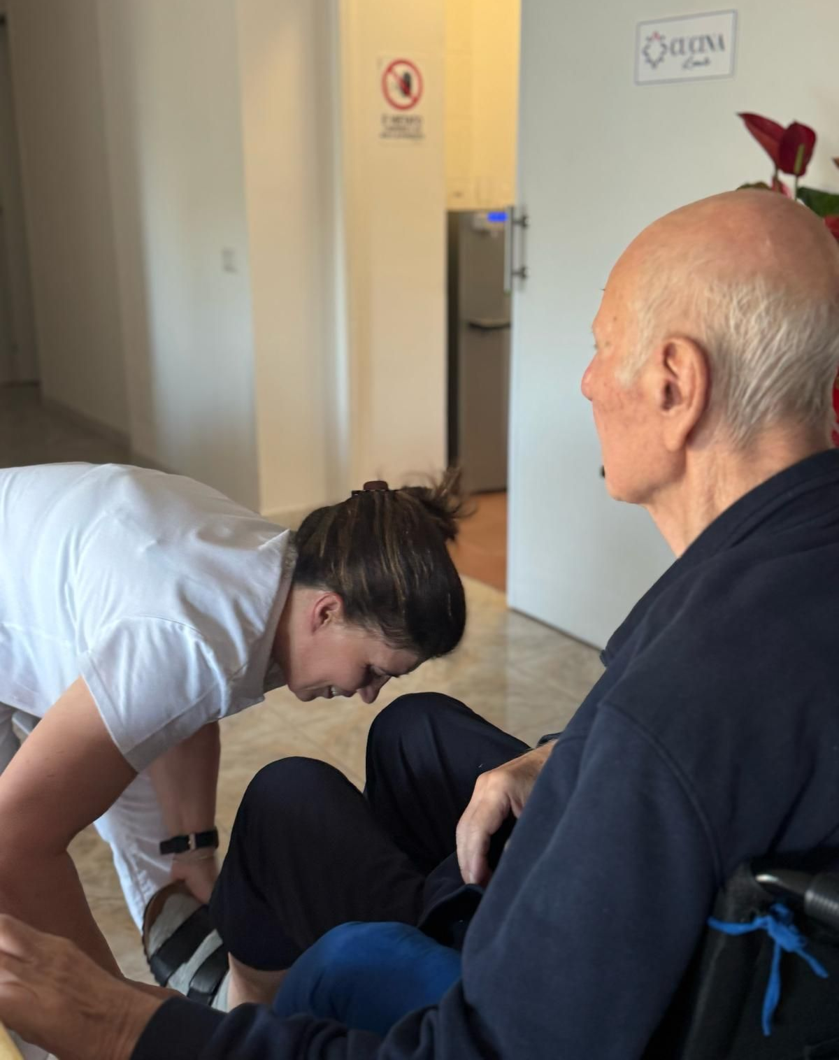 Un operatore sanitario in camice tiene in mano un blocco appunti mentre è in piedi davanti a un gruppo di persone sedute in un ambiente interno luminoso.