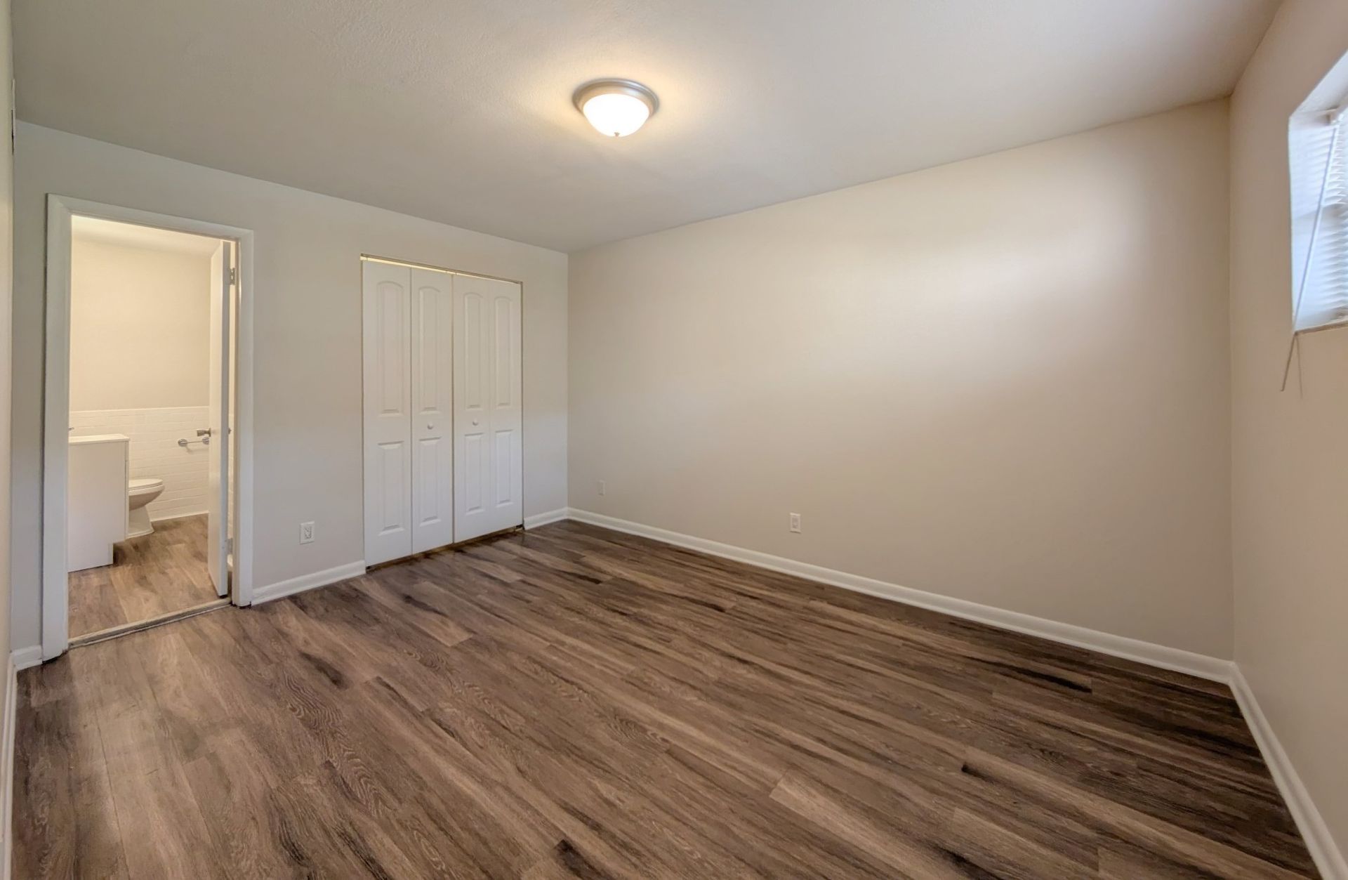 Empty bedroom with closet, bathroom doorway, and wood-look flooring.