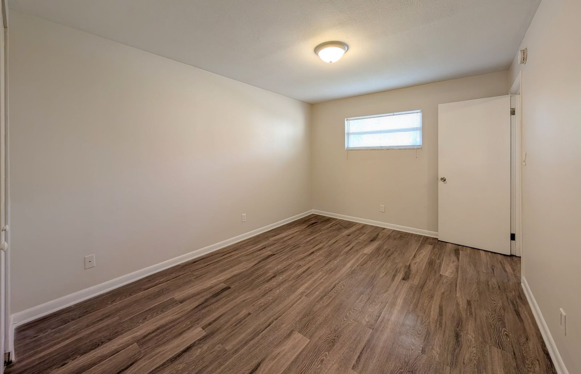 Empty room with wood-look flooring, white walls, small window, and closed door.