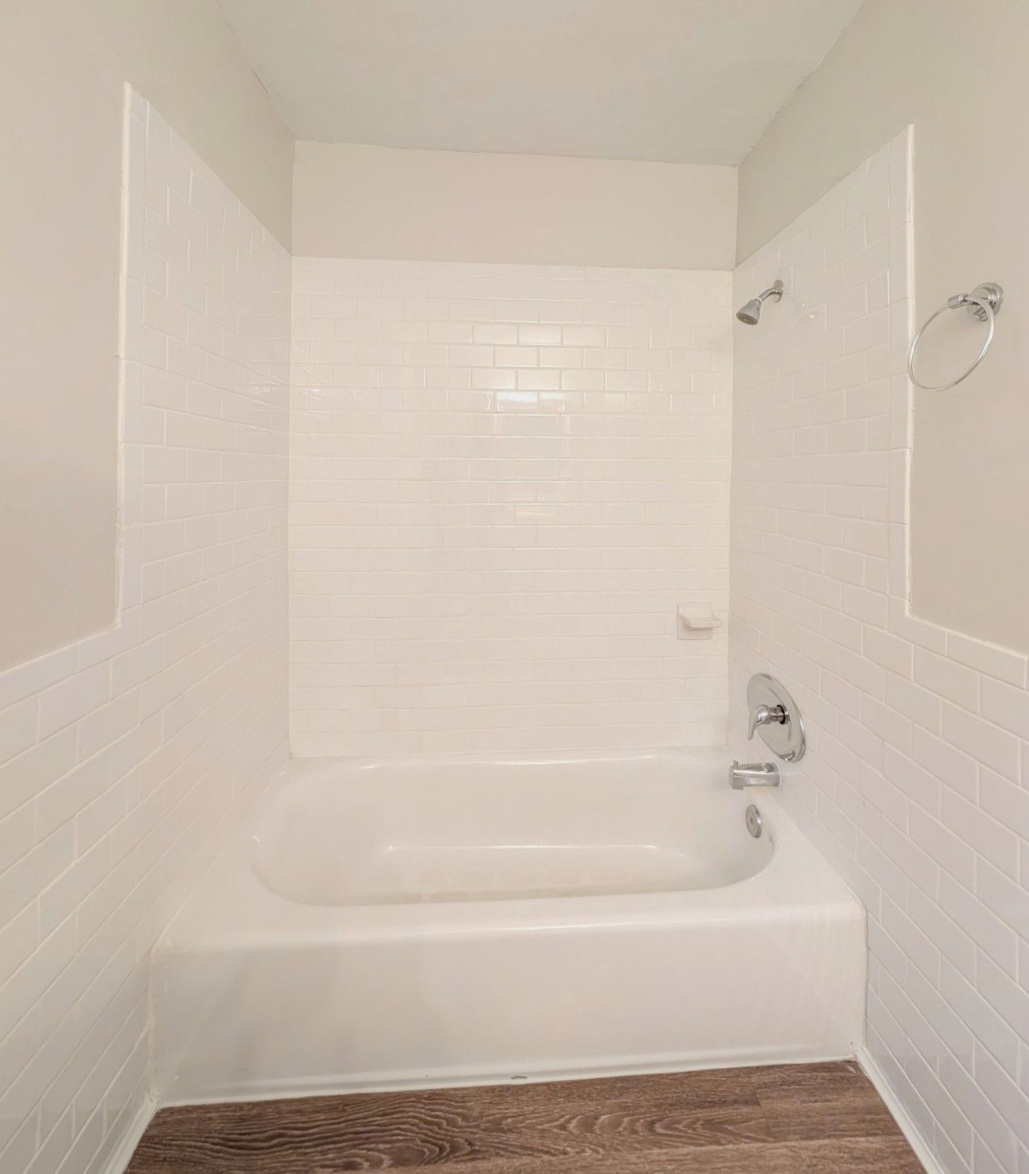 White-tiled shower/bathtub combo with a showerhead and chrome fixtures. The floor is brown, and walls are cream-colored.
