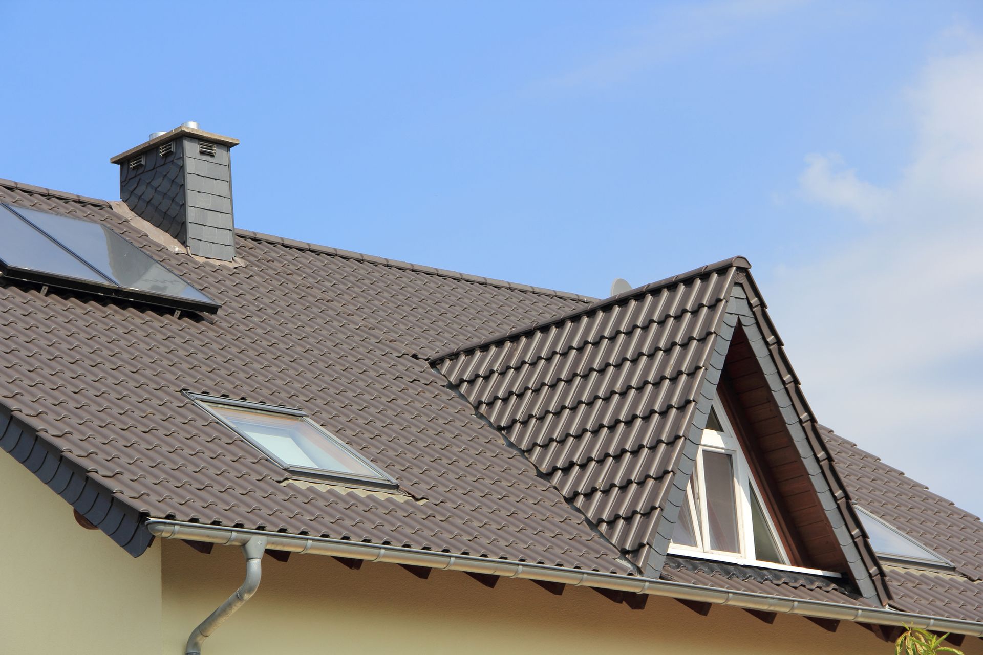 Close-up of a house roof with dark gray tiles, a chimney, and skylights against a blue sky.