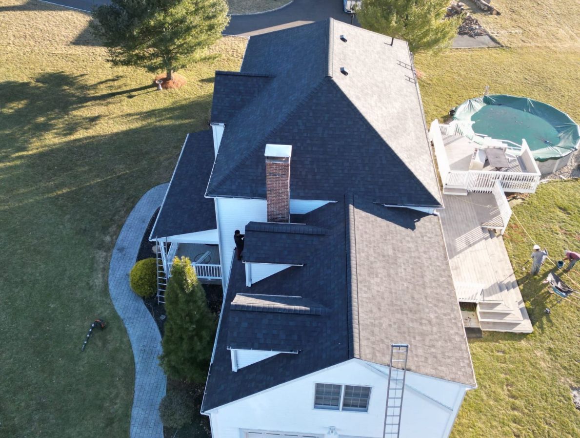 An aerial view of a white house with a dark shingled roof, featuring a brick chimney and two workers on the roof.