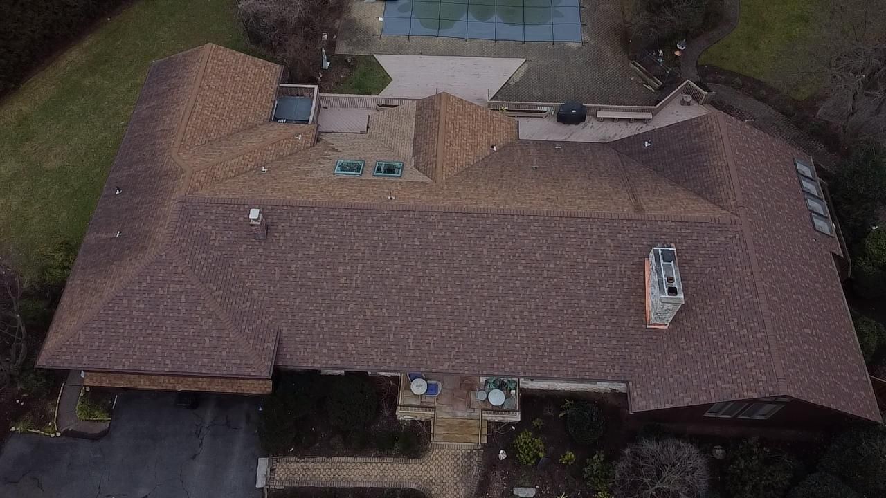 An aerial view of a single-story home with a brown shingled roof, a chimney, and a small pool in the backyard.
