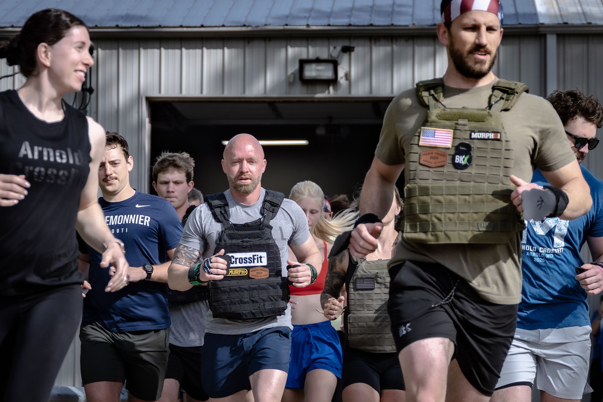 Group of athletes running toward the camera, some wearing weighted vests. Outdoors, sunny. People have focused expressions.