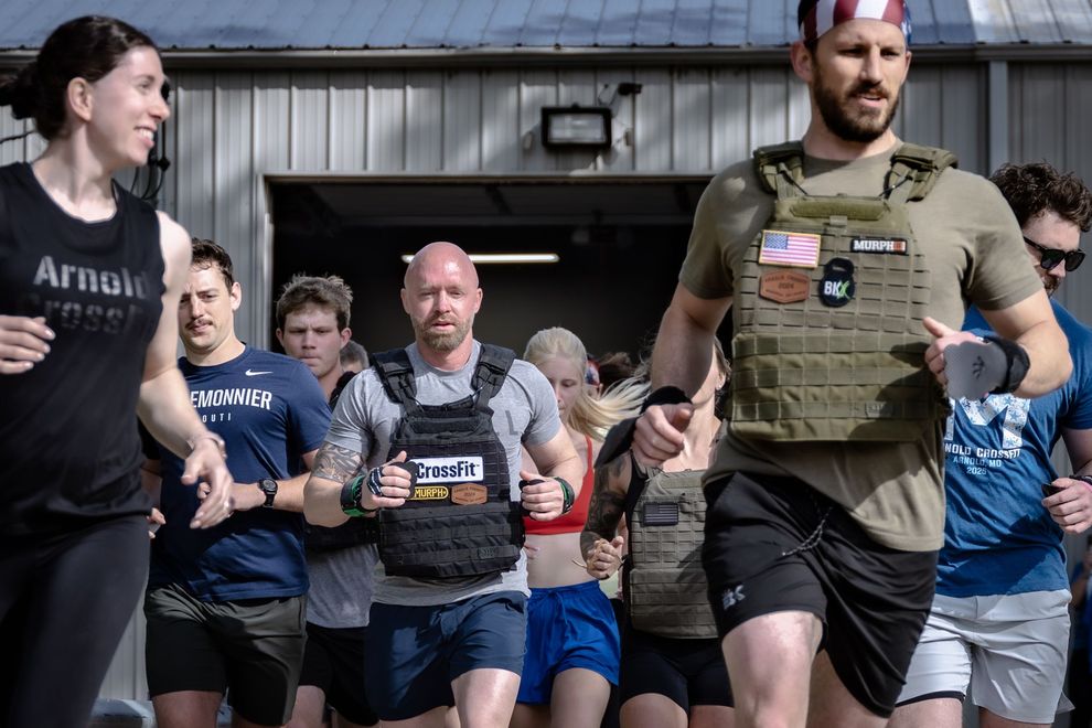 Group of athletes running toward the camera, some wearing weighted vests. Outdoors, sunny. People have focused expressions.