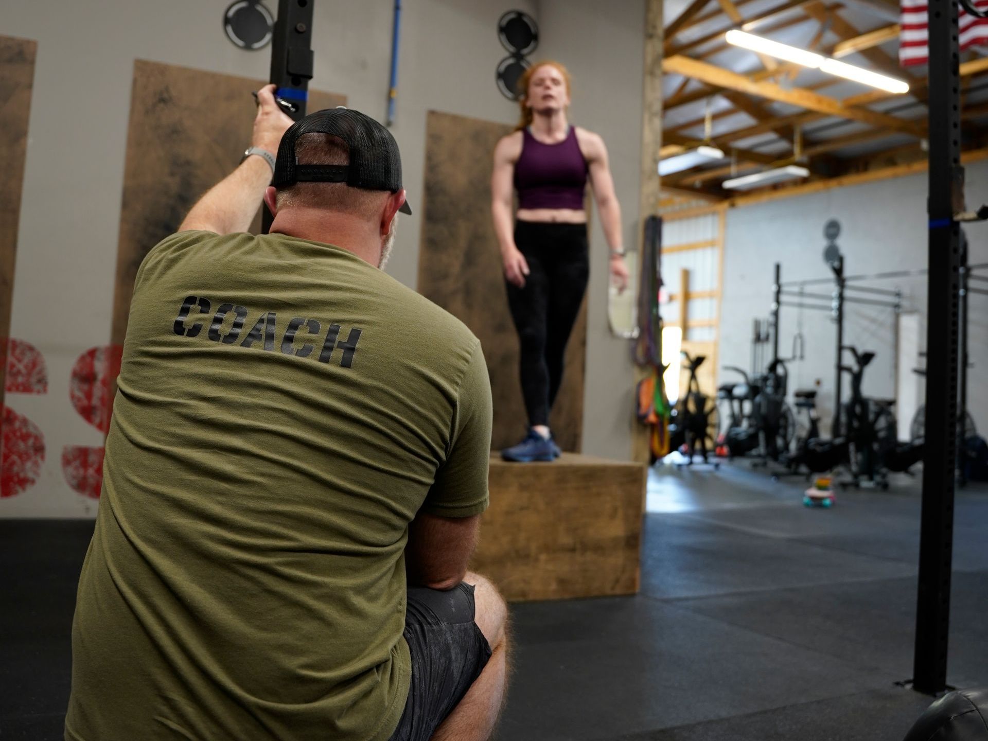 A coach watches a woman perform a box jump in a gym. The coach wears a green shirt with 