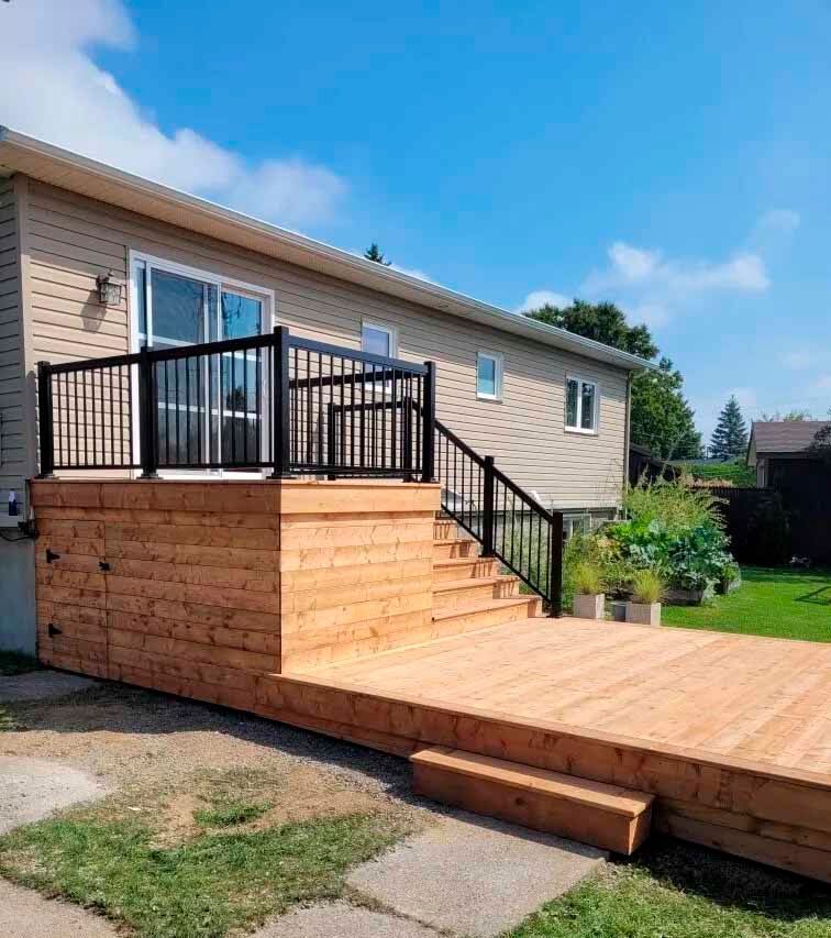 Terrasse en bois avec rambarde noire et marches menant à une maison avec une porte coulissante en verre, par une journée ensoleillée.