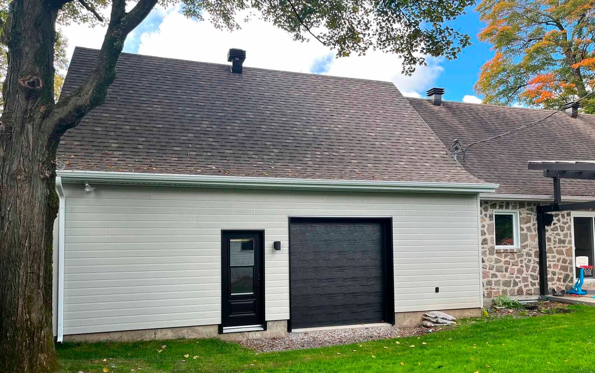 Bâtiment blanc avec porte et porte de garage noires, toit sombre, arbre à gauche et pelouse verte.