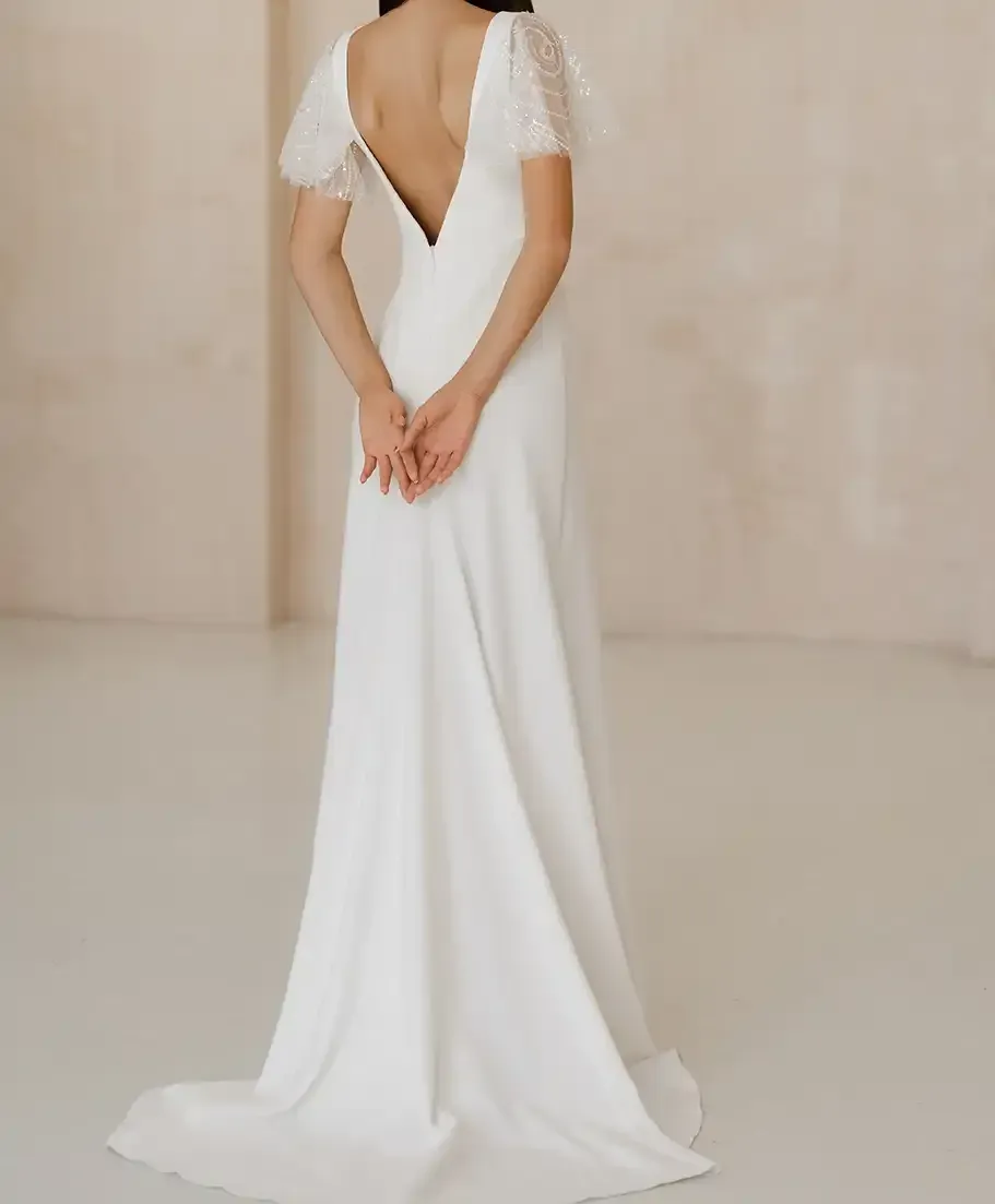 A woman in a white wedding dress is standing in a room with her hands behind her back.