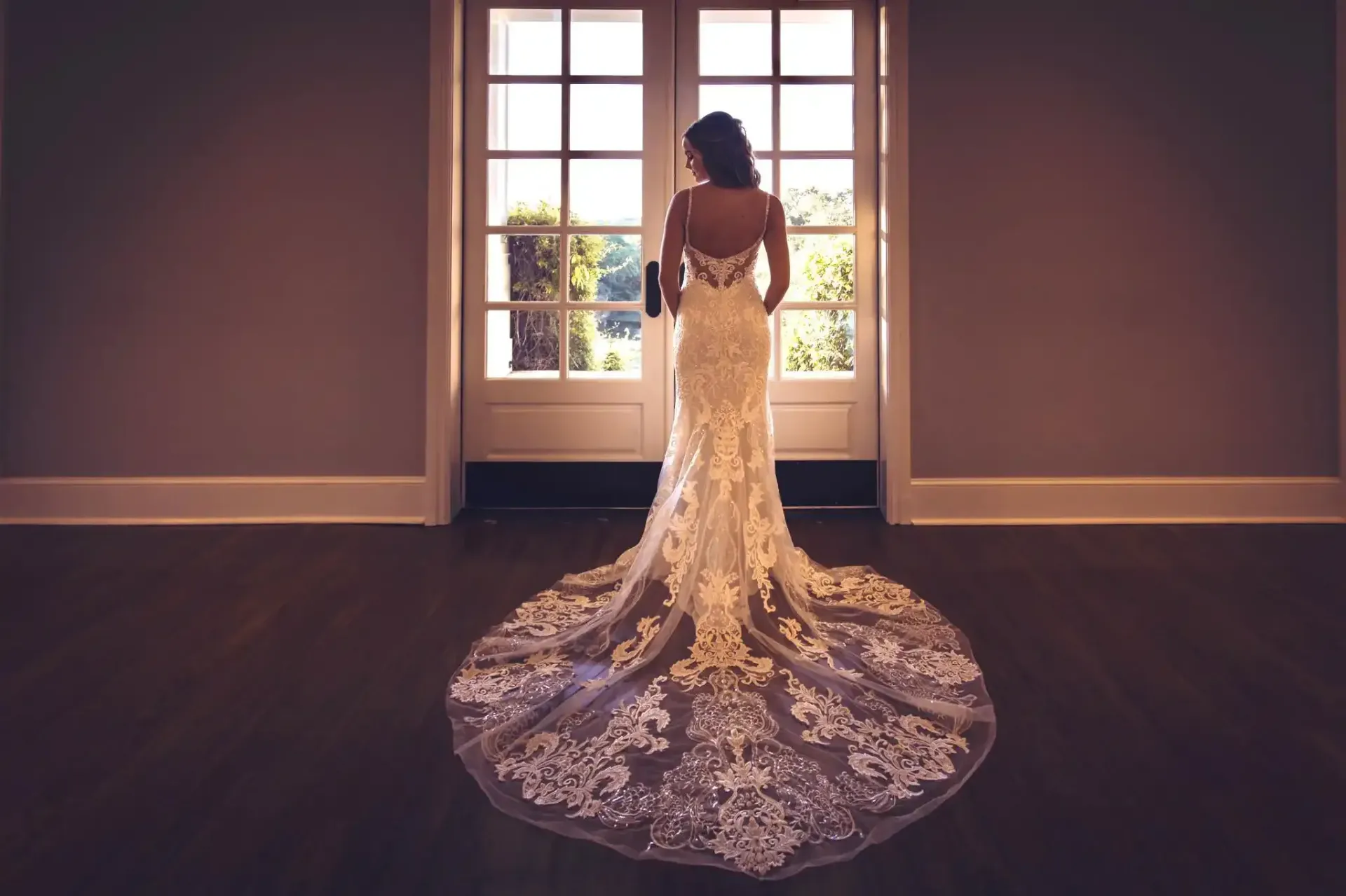 A bride in a long wedding dress is standing in front of a window.